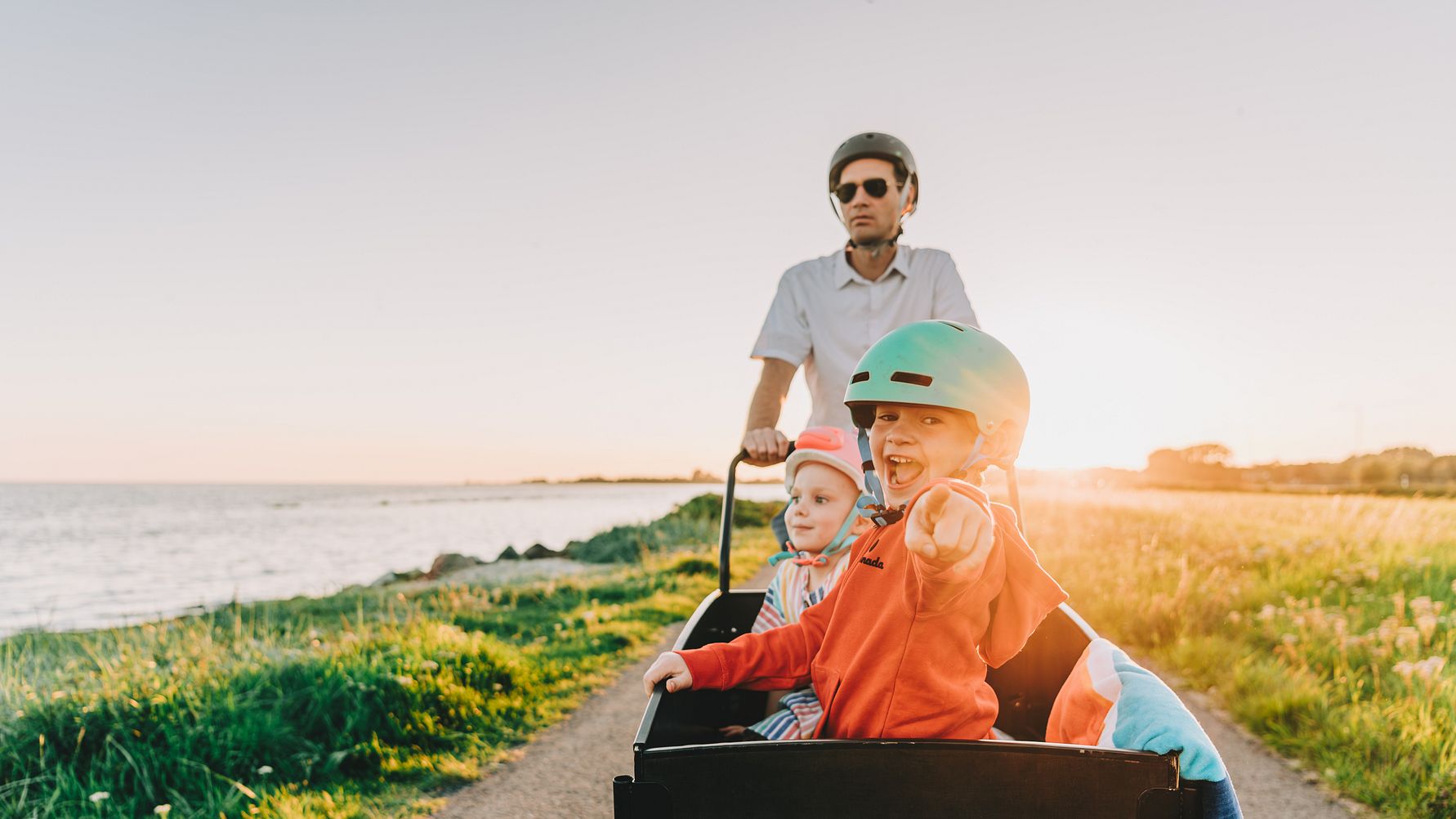 Sommaren på Sydkusten. Pappa och barn cyklar lådcykel med solnedgången i bakgrunden.